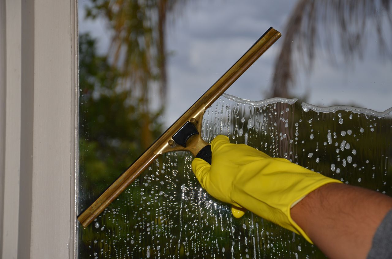 gallery-01 Close-up of hand cleaning window with squeegee outdoors using yellow gloves.